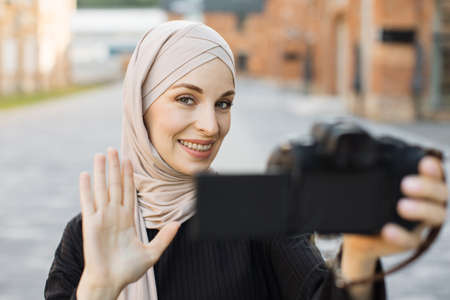 Young muslim woman using a video camera to record journey with smile. Happy and excited 20s arabic girl traveler wear black clothes with hijab, waving to internet audience. Influencer vlogging conceptの写真素材