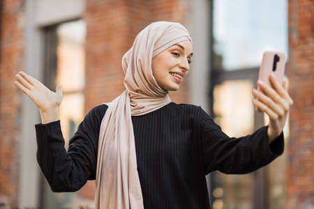 Young smiling happy muslim woman doing video call using smartphone at the city. Arabian woman blogger wearing hijab speaking on the smart phone with her audience walking in the street.の写真素材