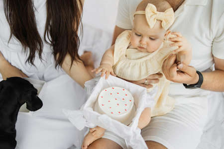 Close up portrait of cheerful young parents embracing their little daughter while sitting together on white blanket with delicious birthday cake. Holidays, celebration and family concert.の写真素材