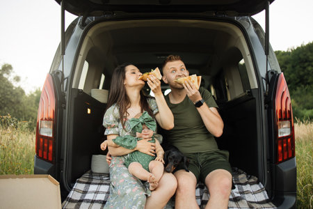 Beautiful married couple with daughter looking at camera while sitting on car trunk with pizza. Celebration, family and happiness concept.の写真素材