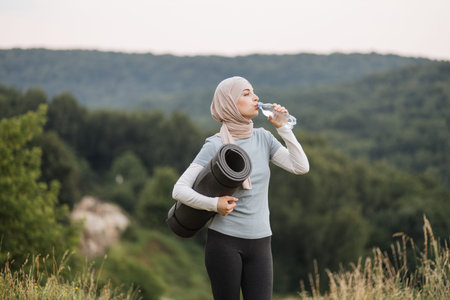 Side view of pretty young woman in hijab and activewear drinking water from bottle while standing at...