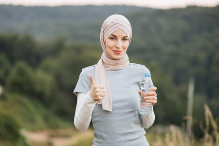 Portrait of charming muslim woman in hijab and sport clothes standing at green park and holding...