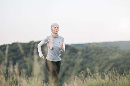 Healthy sport woman in hijab in active clothes enjoying morning run at green park. Muslim woman in hijab spending leisure time for training on fresh air.の写真素材