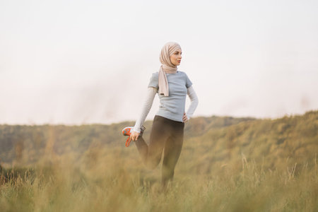 Active woman in sport clothes and hijab stretching legs at summer park. Young arabian girl standing...