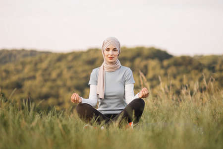 Peaceful young lady in hijab meditating looking at camera in the park. Pretty Muslim woman sitting on mat in yoga pose, finding inner balance.の写真素材