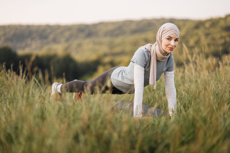 Young muslim woman in hijab and sportswear standing in plank position at green summer park. Sport...