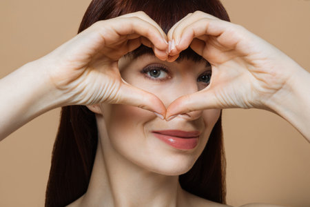 Portrait of positive young lady with healthy smooth skin holding hands in the shape of a heart near...
