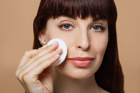 Beautiful red-haired woman, girl cleanses her skin with a cotton pad. Skin care concept, natural beauty. Shot in a studio on a beige background. No makeup.の写真素材