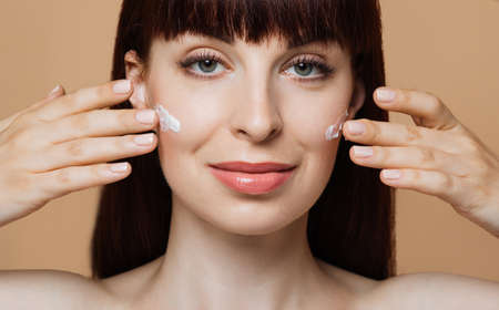Smiling caucasian woman with red hair and healthy skin posing with cream under eyes and naked shoulder on a beige studio wall with free space.の写真素材
