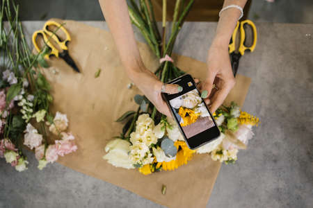 Hands of florist holding smartphone taking picture of bouquet of different flowers on counter. Flat lay, top view mockup. Floristics, business, decoration concept. Close up.の写真素材
