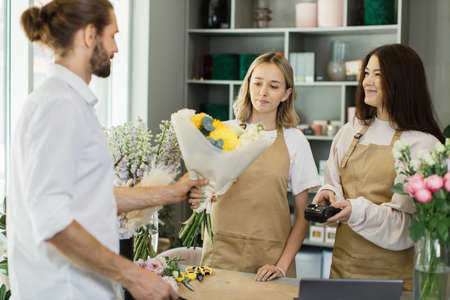 Attractive young bearded man shopping and paying with credit card in flower shop. Male customer paying with card for his purchase bouquet of flowers.の写真素材