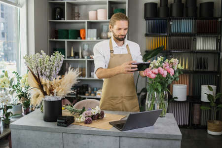 Small business. Attractive young male florist make bouquet and taking photo on smart phone for social media. Flowers delivery, order creation concept.の写真素材