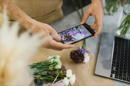 Hands of male florist holding smartphone taking picture of flower on counter. Flat lay, top view mockup. Floristics, business, decoration concept. Close up.の写真素材