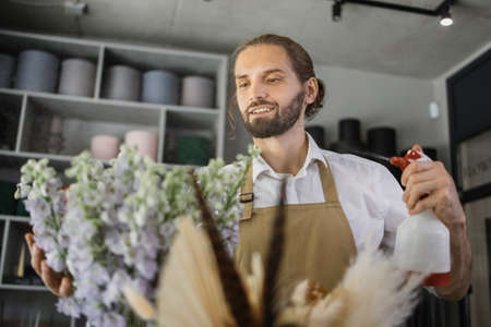 Smiling florist in uniform work in a flower shop behind counter taking care of different varieties of flowers by moisturizing them. Small business concept. Modern loft interior.の写真素材
