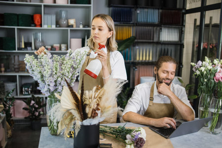 Small family business concept. Two workers caucasian businessman professional designer, florist decorator using laptop at floral store checking inventory of shop and contractor girl waters flowers.の写真素材