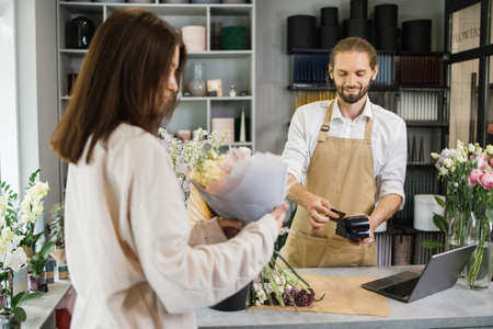 Bearded florist man holding card reader machine at counter with customer paying with credit card. Young florist holding payment machine while female buyer purchase a bunch flower.の写真素材