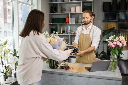 Caucasian female using electronic credit card on her smartphone paying for purchase using payment terminal in flower shop. Client buying bouquet making payment in storeの写真素材