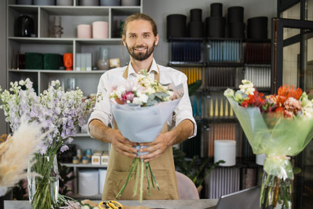 Smiling male florist in uniform work in a flower shop behind counter with different varieties of flowers and huge bouquet in hands. Small business concept. Modern loft interior.の写真素材