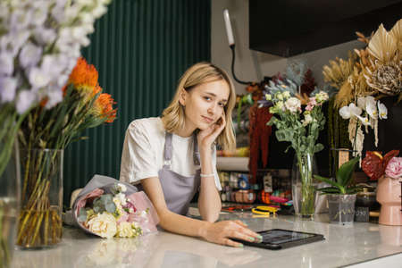 Portrait of attractive woman florist working on tablet at workplace. Concentrated female sitting at counter in floral store and typing on digital tablet.の写真素材