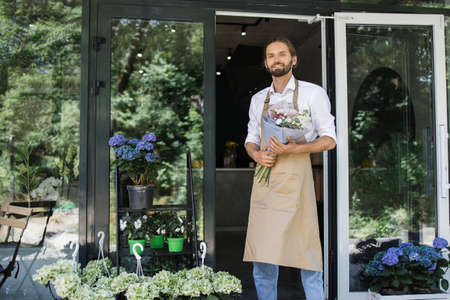 Business man near flower shop door. Photo of handsome man standing in front of flower shop. Male florist outside shop. Woman working at flower shop smiling.の写真素材