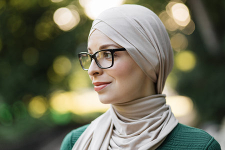 Attractive smiling muslim woman in hijab looking outside standing on green background of city park....