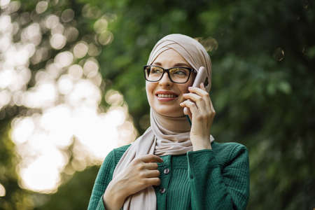 Cheerful young islamic businesswoman talking on mobile phone while walking on city park. Beautiful young woman with hijab working using smartphone during break.の写真素材
