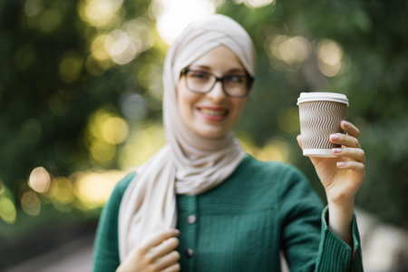Focus on cup of coffee of attractive smiling muslim woman in hijab standing on green background of...