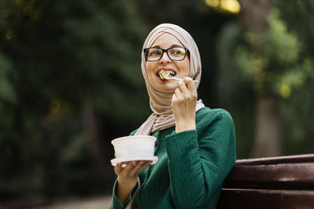 Portrait of a young attractive muslim woman eating salad sitting on the bench on the park. Beautiful young arab female enjoying a fresh salad outdoor.の写真素材