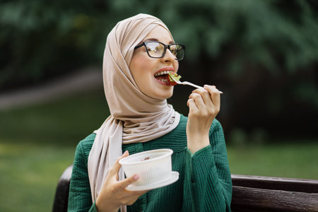 Portrait of happy muslim smiling woman freelancer, manager, business lady, eating fresh vegetable salad during lunch break outdoor in the city park.の写真素材