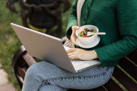 Close up view of hand of businesswoman having a vegetables salad for lunch, healthy eating sitting on bench on the park using modern laptop, lifestyle concept, unrecognizable person.の写真素材