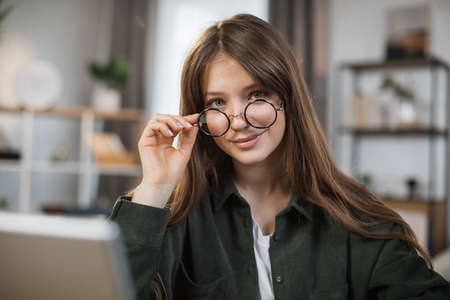 Pretty young caucasian woman with long dark hair sitting at table with modern laptop and touching glasses with hands. Concept of freelance and technology.の写真素材