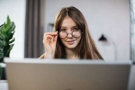 Close up portrait of confident caucasian woman touching glasses with hand looking at camera during video conversation at home. Female freelancer talking and working while sitting at office.の写真素材