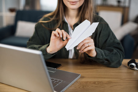 Close up of young woman doing paper plane while sitting at table with laptop and documents. Bored office worker taking fun and playing at workplace.の写真素材