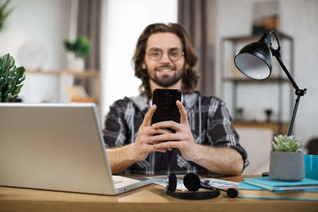 Confident young man writing message during mobile conversation at office. Caucasian male worker with long dark hair sitting at table with opened laptop.の写真素材