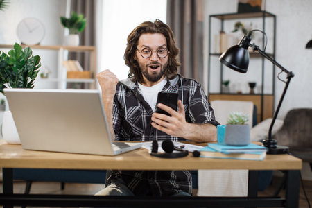 Excited attractive caucasian bearded man sitting at desk and receives good news working on...