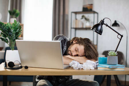 Young frustrated exhausted man laid his head down on the table sitting at desk with pc laptop. Achievement business career concept. Overworked and tired businessman sleeping at work in his office.の写真素材