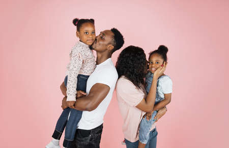 Happy african american woman holding and kissing one daughter, and husband another on pink background, everybody hugging together.の写真素材