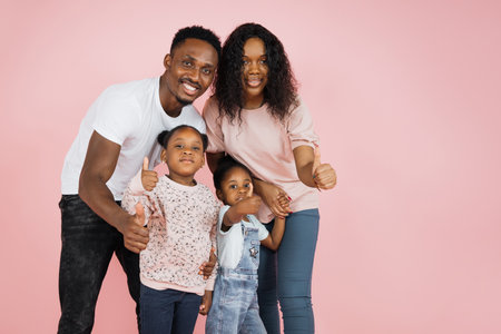 Happy family concept. Young parents and their daughters hugging sitting down on the floor showing thumbs up over pink studio background, free space.の写真素材