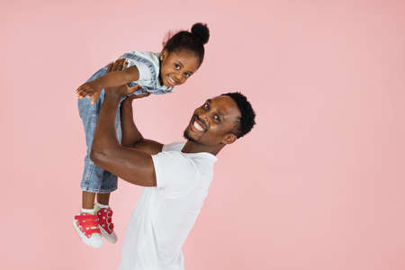 Happy fatherhood. Cheerful african dad holding his cute daughter on hands pretending she are flying, girl spreading hands, imitating plane, pink studio background.の写真素材