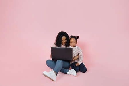 Happy african american mom and little cute daughter spending time together using laptop, sitting together on floor over pink background, copy space.の写真素材