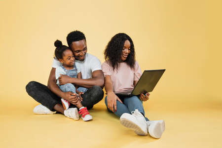 Happy family, technology, communication concept. Young african parents sitting on floor with their daughter and embracing, using laptop for video call, over yellow studio background, free space.の写真素材
