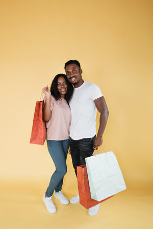 Valentines shopping concept. Smiling black guy and his girlfriend with gift bags on yellow studio...