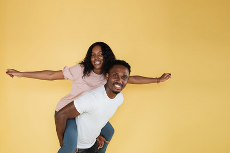 Cheerful smiling young couple, woman with hands aside imitating flying sitting on boyfriend back,...
