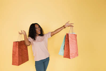 Portrait of positive cheerful afro american girl, addicted shopper, demonstrating a lot of colorful shopping bags to camera, while standing on yellow background, copy space.の写真素材