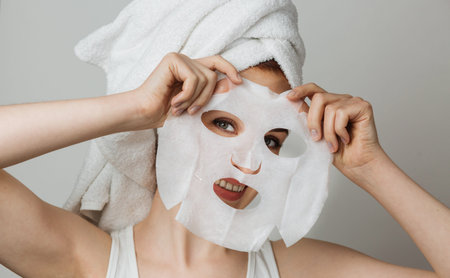 Portrait of beautiful young woman smiling on camera with cotton mask sheet in hands. Isolated over...