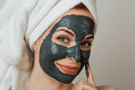 Attractive young woman posing in studio with black clay mask on her face. Caucasian lady in towel...