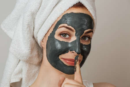 Attractive young lady smiling and looking at camera with black clay mask on her face. Caucasian woman showing sign of silence against grey background.の写真素材
