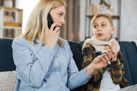 Sad sick teen boy with scarf around neck sitting on sofa at home while his worried young mother talking with doctor by phone with a thermometer after measuring the temperature.の写真素材