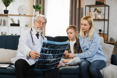Senior male doctor, sick little boy and his mom looking at x-ray scan image during home visit. Young...