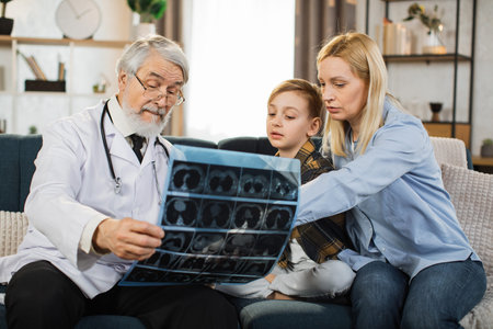 Senior male doctor, sick little boy and his mom looking at x-ray scan image during home visit. Young lady and her child listening their doctor explaining CT scan and talking about the treatment.の写真素材